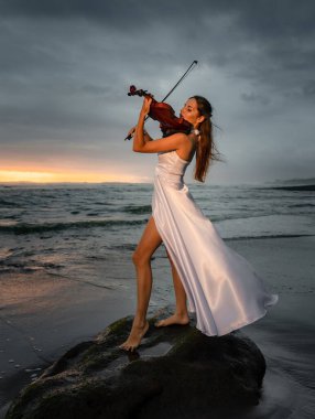 Charming Caucasian woman with violin on the beach. Music and art concept. Slim girl wearing long white dress and playing violin in nature. Sunset time. Cloudy sky. Bali, Indonesia