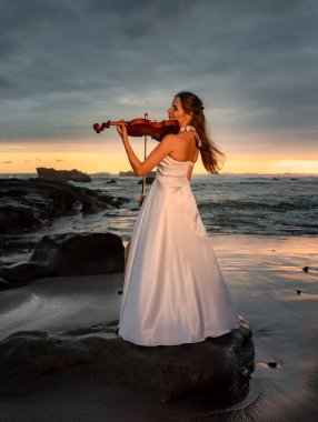 Kumsalda kemanı olan beyaz bir kadın. Müzik ve sanat konsepti. İnce kız uzun beyaz elbise giyiyor ve doğada keman çalıyor. Gün batımı zamanı. Bulutlu gökyüzü. Bali, Endonezya