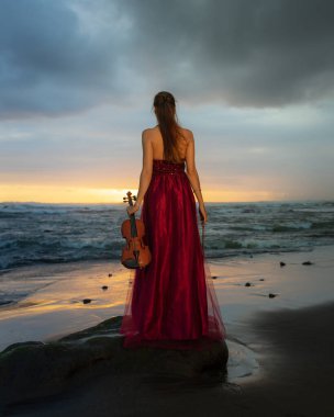 Kumsalda kemanı olan beyaz bir kadın. Müzik ve sanat konsepti. İnce kız uzun kırmızı elbiseler giyiyor ve kemanı doğada tutuyor. Gün batımı zamanı. Bulutlu gökyüzü. Arkadan bak. Bali, Endonezya