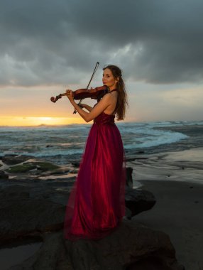 Kumsalda kemanı olan beyaz bir kadın. Müzik ve sanat konsepti. İnce kız uzun kırmızı elbise giyiyor ve doğada keman çalıyor. Gün batımı zamanı. Bulutlu gökyüzü. Bali, Endonezya