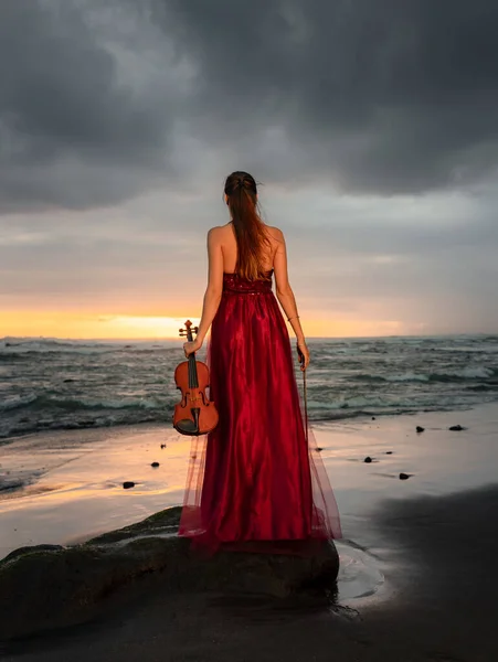 Caucasian woman with violin on the beach. Music and art concept. Slim girl wearing long red dress and holding violin in nature. Sunset time. Cloudy sky. View from back. Bali, Indonesia
