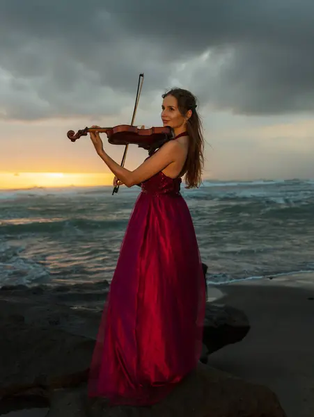 Charming Caucasian woman with violin on the beach. Music and art concept. Slim girl wearing long red dress and playing violin in nature. Sunset time. Cloudy sky. Bali, Indonesia