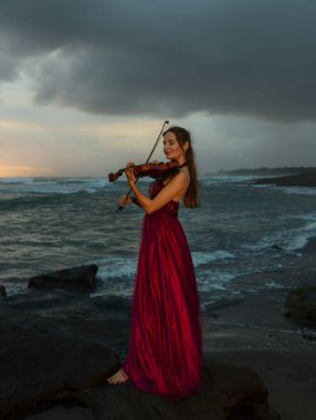 Beautiful Caucasian woman with violin on the beach. Music and art concept. Slim girl wearing long red dress and playing violin in nature. Sunset time. Cloudy sky. Bali, Indonesia