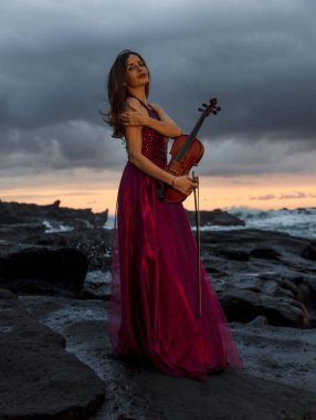 Kumsalda keman çalan çekici beyaz kadın. Müzik ve sanat konsepti. İnce kız uzun kırmızı elbiseler giyiyor ve kemanı doğada tutuyor. Gün batımı zamanı. Bulutlu gökyüzü. Bali, Endonezya