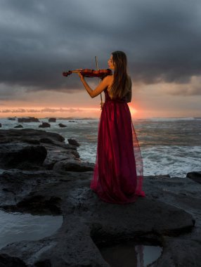 Kumsalda kemanı olan beyaz bir kadın. Müzik ve sanat konsepti. İnce kız uzun kırmızı elbise giyiyor ve doğada keman çalıyor. Gün batımı zamanı. Bulutlu gökyüzü. Arkadan bak. Bali, Endonezya