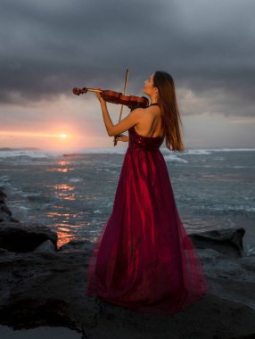 Kumsalda kemanı olan beyaz bir kadın. Müzik ve sanat konsepti. İnce kız uzun kırmızı elbise giyiyor ve doğada keman çalıyor. Gün batımı zamanı. Bulutlu gökyüzü. Bali, Endonezya