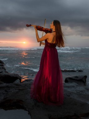 Beautiful Caucasian woman with violin on the beach. Music and art concept. Slim girl wearing long red dress and playing violin in nature. Sunset time. Cloudy sky. Bali, Indonesia