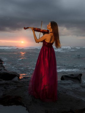 Beautiful Caucasian woman with violin on the beach. Music and art concept. Slim girl wearing long red dress and playing violin in nature. Sunset time. Cloudy sky. Bali, Indonesia