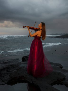Beautiful Caucasian woman with violin on the beach. Music and art concept. Slim girl wearing long red dress and playing violin in nature. Sunset time. Cloudy sky. Bali, Indonesia