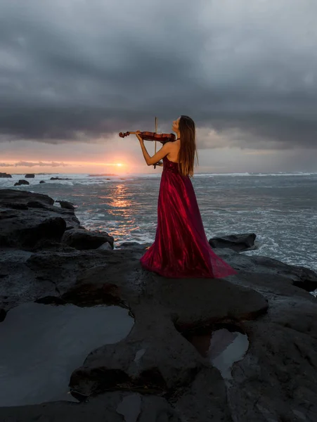 Caucasian woman with violin on the beach. Music and art concept. Slim girl wearing long red dress and playing violin in nature. Sunset time. Cloudy sky. View from back. Bali, Indonesia