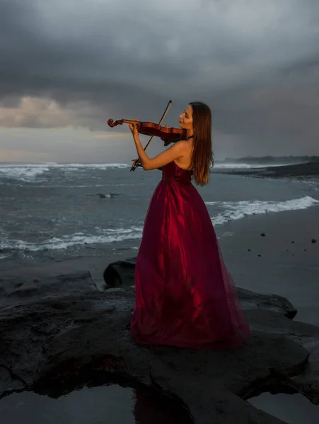 Beautiful Caucasian woman with violin on the beach. Music and art concept. Slim girl wearing long red dress and playing violin in nature. Sunset time. Cloudy sky. Bali, Indonesia