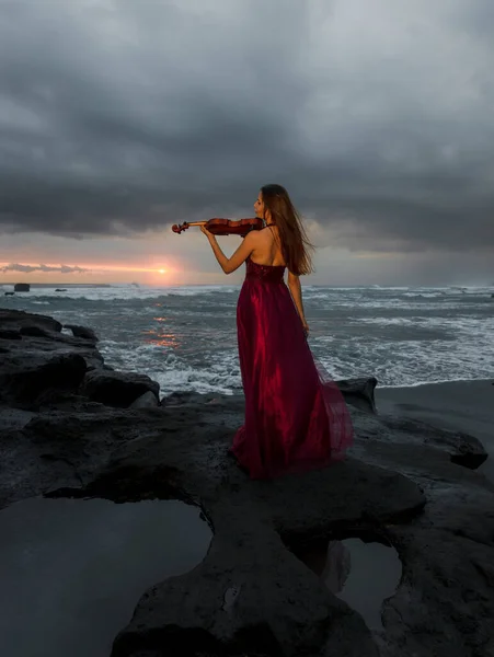 Caucasian woman with violin on the beach. Music and art concept. Slim girl wearing long red dress and playing violin in nature. Sunset time. Cloudy sky. View from back. Bali, Indonesia