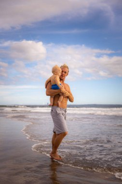 Beyaz bir baba çocuğunu kollarında tutuyor, sahil boyunca yürüyor. Mutlu bir çocukluk. Baba ve oğul yaz tatilini birlikte geçiriyorlar. Dikey yerleşim. Mavi gökyüzü, beyaz bulutlar. Seminyak plajı, Bali