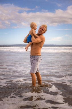 Baba yaz tatillerinin keyfini çıkarıyor, kucağında küçük oğlunu kumlu sahilde havaya kaldırıyor. Aile seyahati ve tatil kavramı. Mavi gökyüzü, beyaz bulutlar. Seminyak Sahili, Bali, Endonezya