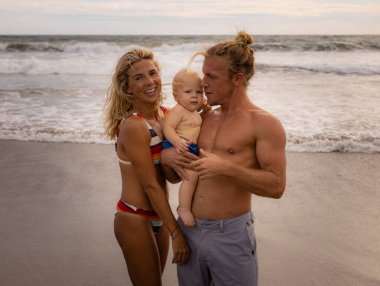 Genç beyaz bir aile kumsalda vakit geçiriyor. Baba kucağında erkek bebek var. Mutlu anne gülümsüyor. Aile ilişkileri. Yaz tatili kavramı. Asya 'ya seyahat. Seminyak Sahili, Bali, Endonezya