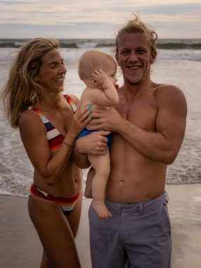 Genç bir aile kumsalda vakit geçiriyor. Baba ve anne kucağında bebek var. Annem ve babam gülümsüyor. Yüzü dönük bir çocuk. Asya 'da yaz tatili. Aile plajı konsepti. Seminyak plajı Bali