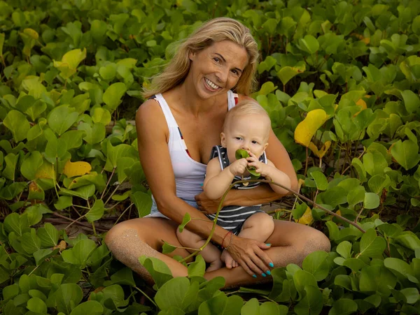 Young Caucasian mother with baby infant boy spending time in nature. Outdoor activities. Mom and baby boy sitting on the ground. Happy child. Background of green leaves. Bali, Indonesia