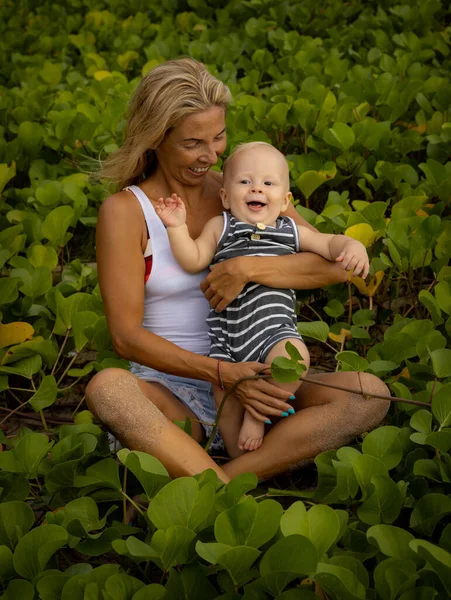Young mother with baby infant boy spending time in nature. Outdoor activities. Mom and baby boy sitting on the ground. Laughing kid. Background of green leaves. Happy childhood. Bali, Indonesia