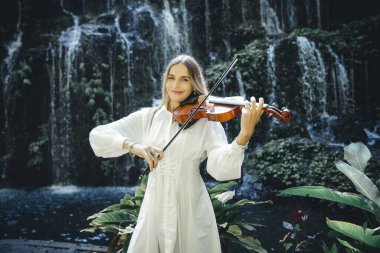 Şelale yakınlarında keman çalan beyaz bir kadın. Müzik ve sanat konsepti. Doğada beyaz elbise giyen zayıf bir kız. Su sıçraması. Banyu Wana Amertha Şelalesi Wanagiri, Bali, Endonezya.