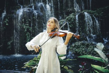 Şelale yakınlarında keman çalan güzel beyaz kadın. Müzik ve sanat konsepti. Doğada beyaz elbise giyen güzel bir kadın. Su sıçraması. Banyu Wana Amertha Şelalesi Wanagiri, Bali, Endonezya.