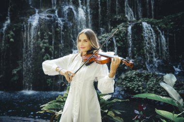 Şelale yakınlarında keman çalan beyaz bir kadın. Müzik ve sanat konsepti. Doğada beyaz elbise giyen zayıf bir kız. Su sıçraması. Banyu Wana Amertha Şelalesi Wanagiri, Bali, Endonezya.
