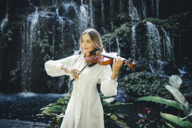 Şelale yakınlarında keman çalan güzel beyaz kadın. Müzik ve sanat konsepti. Doğada beyaz elbise giyen güzel bir kadın. Su sıçraması. Banyu Wana Amertha Şelalesi Wanagiri, Bali, Endonezya.