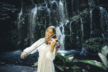 Şelalenin yanında gözleri kapalı keman çalan beyaz bir kadın. Müzik ve sanat konsepti. Zayıf, sarı saçlı, beyaz elbiseli bir kız. Su sıçraması. Banyu Wana Amertha Şelalesi Bali