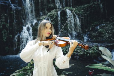 Şelale yakınlarında keman çalan güzel beyaz kadın. Müzik ve sanat konsepti. Doğada beyaz elbise giyen bir kadın. Su sıçraması. Tropik orman. Banyu Wana Amertha Şelalesi, Bali, Endonezya.