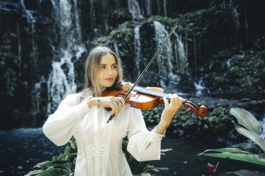 Şelale yakınlarında keman çalan güzel beyaz kadın. Müzik ve sanat konsepti. Doğada beyaz elbise giyen güzel bir kadın. Su sıçraması. Banyu Wana Amertha Şelalesi Wanagiri, Bali, Endonezya.