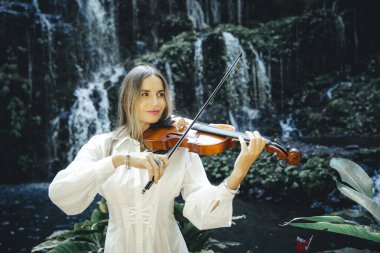 Şelale yakınlarında keman çalan beyaz bir kadın. Müzik ve sanat konsepti. Doğada beyaz elbise giyen bir kadın. Su sıçraması. Tropik orman. Banyu Wana Amertha Şelalesi, Bali, Endonezya.