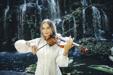 Şelale yakınlarında keman çalan yetenekli beyaz kadın. Müzik ve sanat konsepti. Doğada beyaz elbise giyen güzel bir kız. Su sıçraması. Banyu Wana Amertha Şelalesi Wanagiri, Bali, Endonezya.
