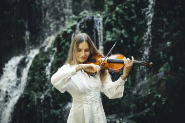 Şelalenin yanında gözleri kapalı keman çalan beyaz bir kadın. Müzik ve sanat konsepti. Zayıf, sarı saçlı, beyaz elbiseli bir kız. Su sıçraması. Banyu Wana Amertha Şelalesi Bali