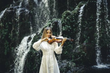 Şelale yakınlarında keman çalan güzel beyaz kadın. Müzik ve sanat konsepti. Doğada beyaz elbise giyen bir kadın. Su sıçraması. Tropik orman. Banyu Wana Amertha Şelalesi, Bali, Endonezya.