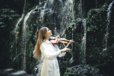 Şelale yakınlarında keman çalan yetenekli beyaz kadın. Müzik ve sanat konsepti. Doğada beyaz elbise giyen çekici bir kadın. Su sıçraması. Kadın yüz profili yan görünümü. Bali 'deki tropikal orman.
