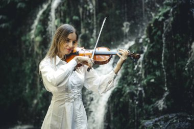 Şelale yakınlarında keman çalan beyaz bir kadın. Müzik ve sanat konsepti. Doğada beyaz elbise giyen bir kadın. Su sıçraması. Tropik orman. Banyu Wana Amertha Şelalesi, Bali, Endonezya.