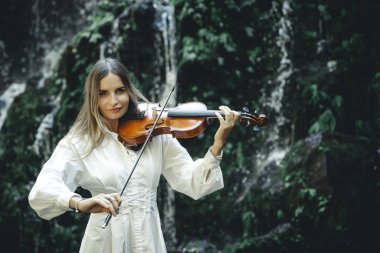 Şelale yakınlarında keman çalan yetenekli beyaz kadın. Müzik ve sanat konsepti. Beyaz elbiseli, uzun sarı saçlı, çekici bir kadın. Su sıçraması. Banyu Wana Amertha Şelalesi Wanagiri, Bali.