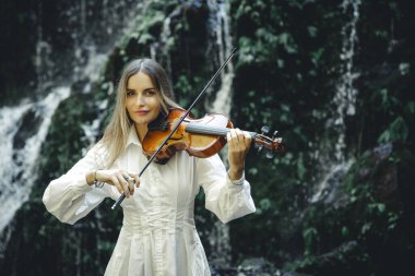 Şelale yakınlarında keman çalan güzel beyaz kadın. Müzik ve sanat konsepti. Beyaz elbiseli, uzun sarı saçlı, çekici bir kadın. Su sıçraması. Banyu Wana Amertha Şelalesi Wanagiri, Bali.