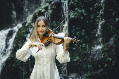 Şelale yakınlarında keman çalan güzel beyaz kadın. Müzik ve sanat konsepti. Beyaz elbise giyen, uzun sarı saçlı bir kadın. Su sıçraması. Banyu Wana Amertha Şelalesi Wanagiri, Bali. Boşluğu kopyala.