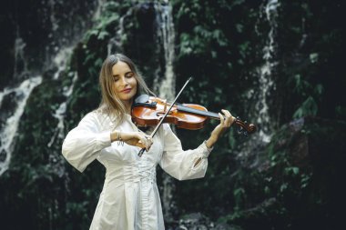 Şelalenin yanında keman çalan güzel beyaz kadın. Müzik, sanat konsepti. Beyaz elbiseli, sarı saçlı, çekici bir kız. Su sıçraması. Banyu Wana Amertha şelalesi. Boşluğu kopyala.