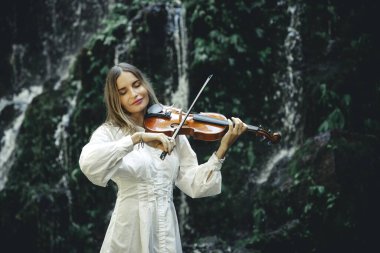 Şelalenin yanında gözleri kapalı keman çalan beyaz bir kadın. Müzik, sanat konsepti. Beyaz elbise giyen, uzun sarı saçlı bir kadın. Su sıçraması. Banyu Wana Amertha şelalesi. Boşluğu kopyala.