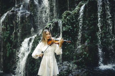 Şelale yakınlarında keman çalan beyaz bir kadın. Müzik ve sanat konsepti. Doğada beyaz elbise giyen bir kadın. Su sıçraması. Tropik orman. Banyu Wana Amertha şelalesi, Bali. Boşluğu kopyala.