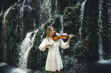 Şelale yakınlarında keman çalan güzel beyaz kadın. Müzik ve sanat konsepti. Beyaz elbiseli, uzun sarı saçlı, çekici bir kadın. Su sıçraması. Banyu Wana Amertha şelalesi, Bali. Boşluğu kopyala.