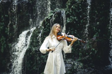 Şelale yakınlarında keman çalan güzel beyaz kadın. Müzik ve sanat konsepti. Doğada beyaz elbise giyen çekici bir kadın. Su sıçraması. Banyu Wana Amertha Şelalesi Wanagiri, Bali, Endonezya.