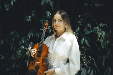 Tropikal ormanda keman tutan güzel beyaz kadın portresi. Müzik ve sanat konsepti. Beyaz elbiseli kız. Açık hava konsepti. Yeşilli yeşil tropikal yaprakların arka planı. Bali, Endonezya.