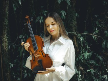 Tropikal ormanda keman tutan beyaz bir kadının portresi. Kapalı gözler. Müzik ve sanat konsepti. Beyaz elbiseli bir kadın. Yeşilli yeşil tropikal yaprakların arka planı. Bali, Endonezya.