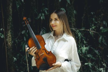 Tropikal ormanda keman tutan gülümseyen beyaz kadın portresi. Müzik ve sanat konsepti. Beyaz elbise giyen, uzun sarı saçlı bir kadın. Yeşillik tropik yaprakların arka planı. Bali, Endonezya.