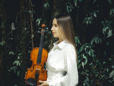 Tropikal ormanda keman tutan güzel beyaz kadın portresi. Kadın yüz profili yan görünümü. Müzik ve sanat konsepti. Beyaz elbiseli kız. Yeşilli yeşil tropikal yaprakların arka planı. Bali.