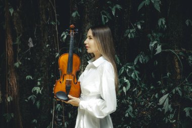 Tropikal ormanda keman tutan beyaz bir kadının portresi. Kadın yüz profili yan görünümü. Müzik ve sanat konsepti. Beyaz elbiseli bir kadın. Yeşilli yeşil tropikal yaprakların arka planı. Bali