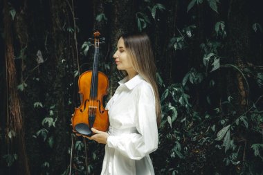 Tropikal ormanda keman tutan beyaz bir kadının portresi. Kadın yüz profili yan görünümü. Müzik ve sanat konsepti. Beyaz elbiseli bir kadın. Yeşillik tropik yaprakların arka planı. Boşluğu kopyala.