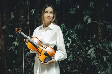 Tropikal ormanda keman tutan beyaz kadın portresi. Müzik ve sanat konsepti. Beyaz elbise giyen, uzun sarı saçlı bir kadın. Yeşillik tropik yaprakların arka planı. Bali, Endonezya. Boşluğu kopyala.